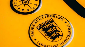 A close-up of a yellow vehicle registration plate displaying black symbols and text. The upper section features a circular inspection sticker with numerical markings from 1 to 12 and a highlighted number 10 in the center. Below, a circular emblem contains the text 'Baden-Württemberg' and a stylized coat of arms with lions.