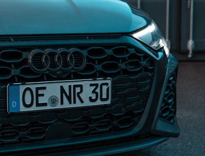 The image features the front end of a car with a visible license plate reading 'OE NR 30'. The car has a sleek black grille and is parked in front of what appears to be an industrial or shipping container backdrop.