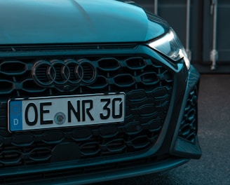 The image features the front end of a car with a visible license plate reading 'OE NR 30'. The car has a sleek black grille and is parked in front of what appears to be an industrial or shipping container backdrop.