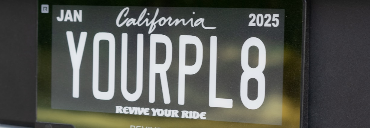 Close-up view of a car's rear license plate with a custom plate that reads 'YOURPL8'. The license plate is issued from California, valid until January 2025. The car's paint appears to be blue.