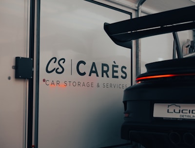 A garage with signage displaying the words 'CARÈS Car Storage & Services' on a glass door. Part of a sports car with a large rear spoiler, visible from the back, occupies the right side of the image, with 'LUCID Detailing' on its license plate.