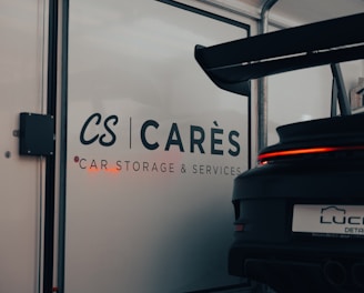 A garage with signage displaying the words 'CARÈS Car Storage & Services' on a glass door. Part of a sports car with a large rear spoiler, visible from the back, occupies the right side of the image, with 'LUCID Detailing' on its license plate.