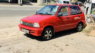 A small red car is parked on a brick-paved area near a road. The car has a license plate reading '302-6413' and black, five-spoke wheels. In the background, there is a service station with signs for 'xpress lube' and 'Havoline', along with trees and vegetation visible in the vicinity.